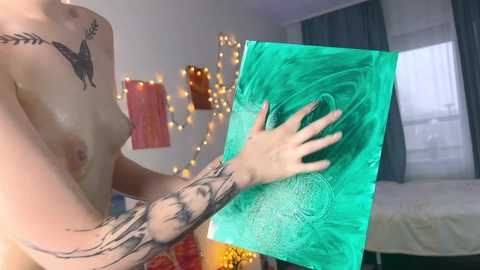 Media: Video of a topless woman with fair skin and medium-sized breasts, tattoos on her arm and shoulder, holding a green, marbled paper against a background of fairy lights, a bed, and a window.