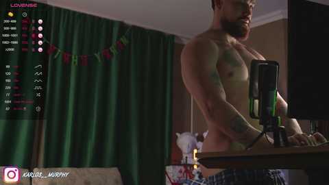 Media: Video of a shirtless, muscular, bearded man in underwear, sitting at a desk with a computer and microphone, in a dimly-lit room with green curtains and Christmas decorations.