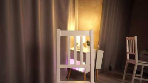 Media: A video of a white wooden chair with vertical slats, positioned in a dimly lit room with beige curtains and a warm, golden light emanating from a nearby lamp, casting a soft glow.