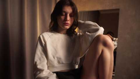 Media: Video of a young woman with fair skin, long dark hair, and a contemplative expression, sitting with legs bent and resting one arm on her knee. She wears a white sweatshirt and dark shorts. The background includes a mirror and beige curtains.