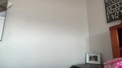 Media: Video of a minimalist bedroom corner with a white wall, wooden headboard, framed photo, and motivational black-and-white sign on the right.