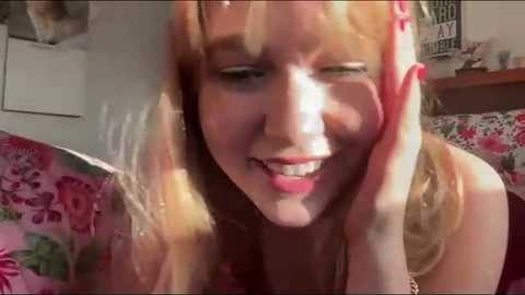 Media: A video of a blonde woman with fair skin, wearing a pink floral dress, smiling widely while touching her cheek, in a bedroom with a pink floral bedspread and a framed \"LOVE\" sign.