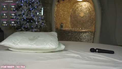 Media: Video of a bed with a white pillow and a black vibrator on the white sheets, with a snowy Christmas tree and fireplace in the background, set in a cozy, warmly lit room.