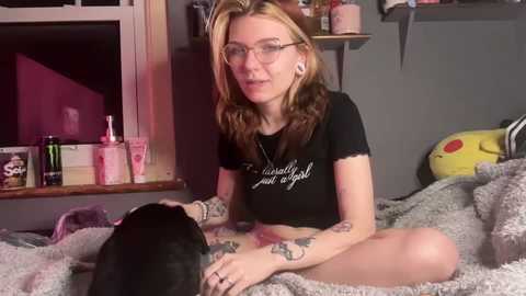 Media: Video of a young woman with fair skin, light brown hair, and glasses, wearing a black crop top and sitting on a bed with a gray blanket. Background shows a cluttered shelf with various items.