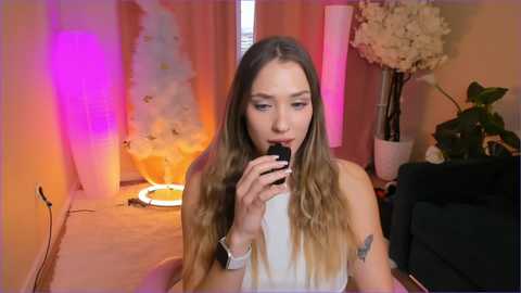 Media: A young woman with long, wavy hair, wearing a white top, sits in a living room with soft lighting, drinking from a black cup, tattoos on her arm, flowers, and furniture in the background.