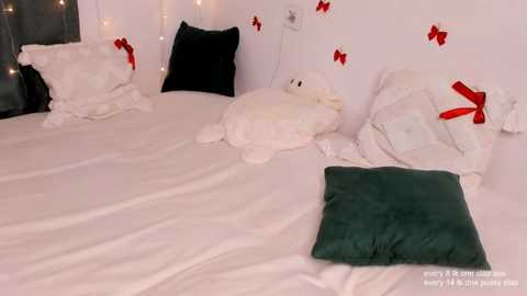 Media: Video of a cozy, white bed with plush toys (penguin, turtle, and others), green and white pillows, and red ribbon decorations on a white wall, creating a festive, holiday-themed atmosphere.