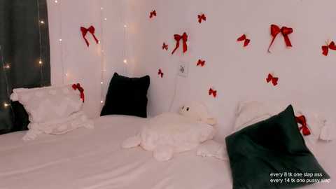 Video of a cozy bedroom with white bedding, plush toys, and festive red bows on the wall, illuminated by fairy lights.