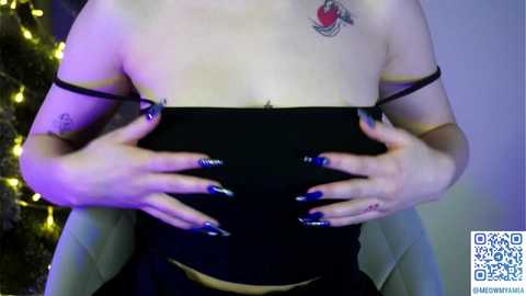 A video shows a light-skinned person with a tattoo of a red heart on their left shoulder, wearing a black top and blue nail polish. They hold the top with both hands, revealing their bare chest. A Christmas tree with yellow lights is visible in the background.