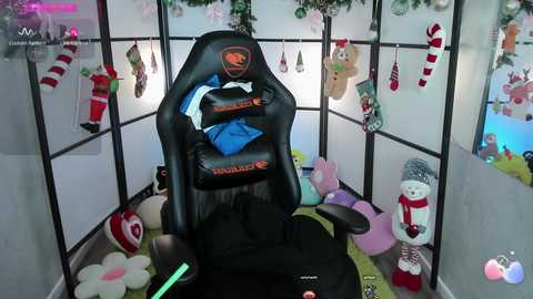 Media: Video of a black gaming chair with a blue pillow, surrounded by plush toys, festive stockings, and a snowman, in a cozy, Christmas-themed room with a white screen backdrop.