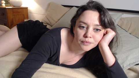 Media: Video of a fair-skinned woman with long dark hair, wearing a black top and black shorts, lying on a beige bedspread in a cozy bedroom with wooden furniture and a soft lamp.