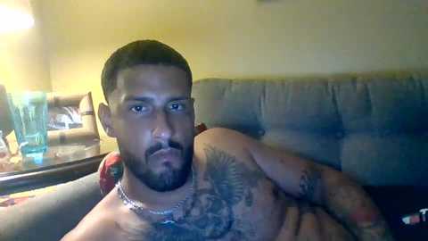 Media: Video of a shirtless, bearded Latino man with a tattooed chest, wearing a silver chain necklace, reclining on a couch with a beige cushion. Background includes a blue glass and a beige armchair.