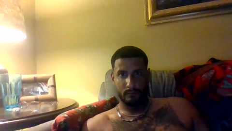 Media: Video of a bearded man with a tattoo on his chest, sitting on a bed, wearing a necklace, in a dimly lit room with a lamp and framed art on the wall.