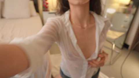 Media: A blurred video of a woman in a semi-transparent white blouse, revealing her cleavage, standing in a dimly-lit bedroom with a bed, nightstand, and lamp visible.