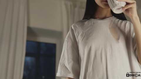 Media: Video of a young East Asian woman with long black hair, wearing a loose-fitting, white T-shirt with a large, visible wet stain on the left side, holding a glass. The background shows a dimly lit room with white curtains and a window.