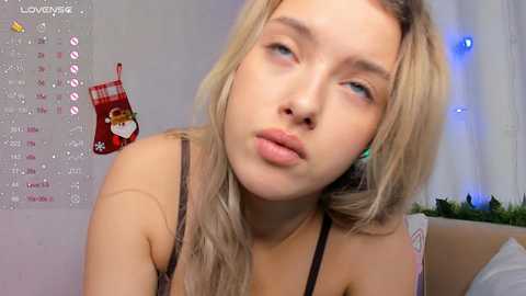 Media: Video of a young, fair-skinned, blonde woman with blue eyes, wearing a black bra, pouting in a cozy, Christmas-themed room with a calendar and festive decorations.