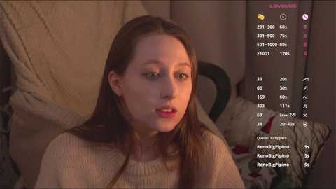 Media: Video of a young woman with fair skin and long brown hair, wearing a beige sweater, sitting on a patterned cushion. Background includes a soft, textured fabric and a partially visible chair.