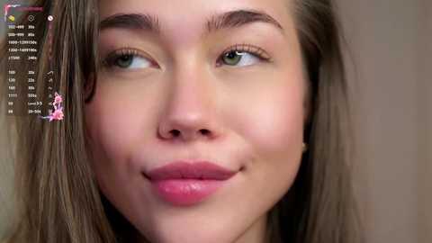 Video of a young Caucasian woman with fair skin, green eyes, and light brown hair, smiling subtly. Background is a blurred, neutral tone, and a digital overlay shows a pink heart and text in the top left corner.