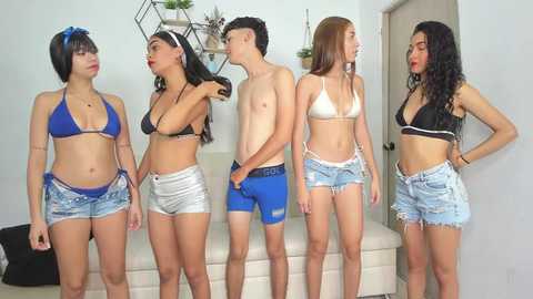 Media: Video of four young women in bikinis and denim shorts, one man in swim trunks, standing in a modern, minimalist room with white walls and a beige couch.