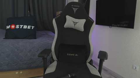 Media: Video of a black gaming chair with a gray pillow, placed in a dimly lit bedroom with a bed, dark curtains, and a mounted TV.