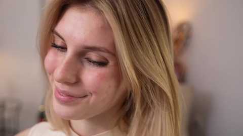 Video of a young Caucasian woman with fair skin and straight, blonde hair. She has closed eyes, a soft smile, and wears a white top. The blurred background shows a beige wall.