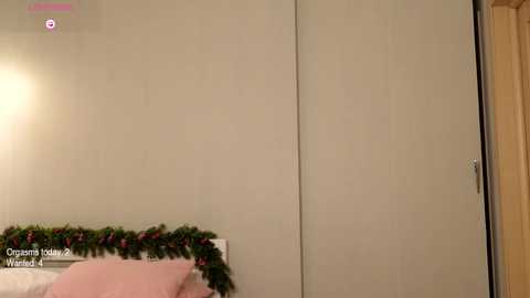 Media: Video of a minimalist, beige-walled bedroom with a closed white wardrobe, a Christmas garland, and a pink throw pillow on a bed.