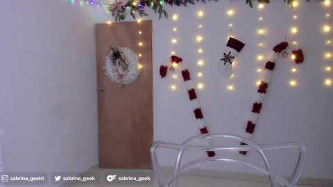 Media: Video of a minimalist Christmas scene: white wall, wooden door with wreath, red and white candy canes, white chair, string lights, and a small tree in the background.