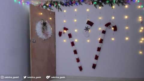 Media: Video of a festive room with a white wall adorned with a garland of multicolored lights, a red and white candy cane, and a snowflake decoration. A beige door with a wreath and a \"Welcome\" sign is visible.