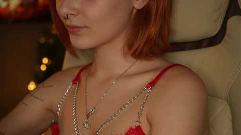 Media: Video of a fair-skinned woman with short red hair, wearing a red lace bra and multiple silver chain necklaces, seated in a beige leather chair.