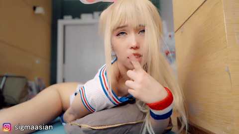 Media: Video of a young, fair-skinned woman with long, blonde hair and pink bunny ears, wearing a white crop top and blue shorts, with a seductive expression, in a dimly lit hallway.