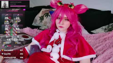 Media: Video of a fair-skinned woman with long, bright pink hair in a Santa costume, lying on a bed with a pink blanket. Background includes a screen with a \"Love Live!\" game and a plush toy.