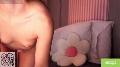 Media: Video of a topless, light-skinned woman with small breasts, sitting on a bed with a large, white daisy pillow. The background features pink and white striped wallpaper.