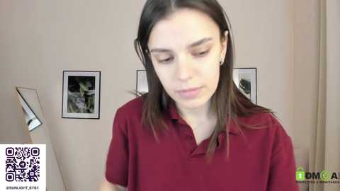 Media: A video of a young woman with shoulder-length brown hair, wearing a maroon polo shirt, looking downward in a well-lit room with beige walls and framed art.