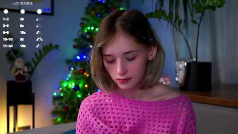 Video of a young, light-skinned woman with short blonde hair wearing a pink knitted sweater, standing in a dimly-lit room with a decorated Christmas tree and potted plants.