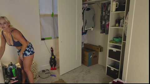 Media: Video of a messy bedroom with a blonde woman in a blue and white patterned bra and shorts, leaning on a white wall, surrounded by clutter.