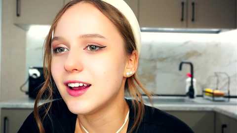 Media: Video of a young woman with fair skin, brown hair, and green eyes, wearing a white headscarf, black top, and pearl necklace, in a modern kitchen with white cabinets, stainless steel sink, and a black kettle.