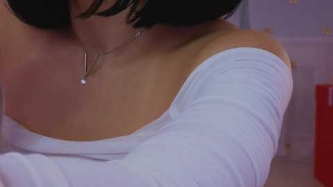 Media: Video of a woman with a short black bob haircut, wearing a white off-shoulder top, and a delicate necklace with a small pendant, indoors with soft purple and red background.