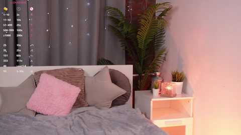 Media: Video of a cozy bedroom with a white bed, gray blanket, and pink pillow. Background includes a potted plant, white shelves, and a window with gray curtains.