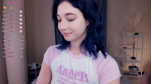 Media: A video of a young woman with shoulder-length dark hair wearing a pink Angels shirt, standing in a dimly lit room with a calendar, shelves, and a lamp.