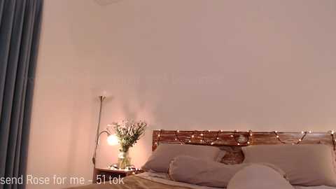 Media: Video of a minimalist bedroom featuring a wooden bed with white pillows, a small side table with a vase of flowers, and a blue curtain on the left.