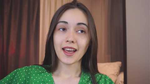 Media: Video of a young woman with long, straight brown hair and fair skin, smiling with slightly parted lips. She wears a green polka-dot top and is indoors with beige curtains and a bed visible in the background.