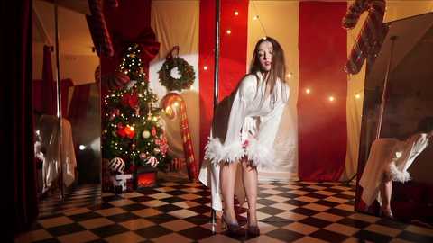 Media: Video of a woman in a white, fluffy robe, standing on a black-and-white checkered floor, near a decorated Christmas tree and festive red curtains in a cozy, warmly lit room.