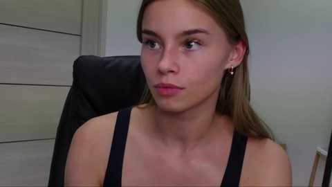 Media: Video of a young woman with light skin, brown hair, and green eyes, wearing a black tank top, sitting in a black leather chair. She has a serious expression. Background shows a wooden door and light-colored walls.