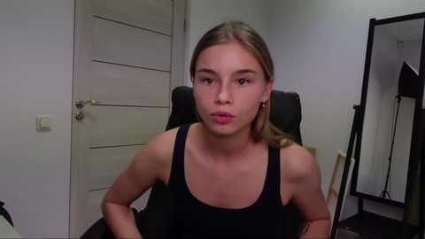 Media: Video of a young woman with light skin and straight, shoulder-length blonde hair, wearing a black tank top, sitting in a black office chair in a minimalist room with white walls and a closed door.