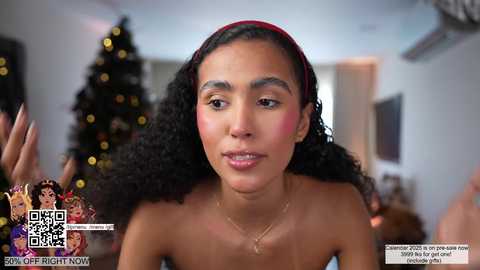 Media: Video of a young Black woman with curly hair, wearing a red headband and makeup, smiling, in a festive indoor setting with a decorated Christmas tree.