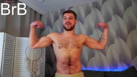 Media: Video of a muscular, shirtless man with a beard and short hair flexing his biceps. Background features a gray, leaf-patterned wall and a white screen with a dreamcatcher.