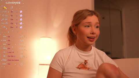 Media: A video of a young, fair-skinned, blonde-haired girl in a white T-shirt with a cartoon dog, smiling in a warmly lit room with a lamp and wall calendar.