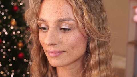 Media: Video of a Caucasian woman with wavy blonde hair, light freckles, and light makeup, looking downward. Background features a blurred, festive Christmas tree with multicolored lights and ornaments.