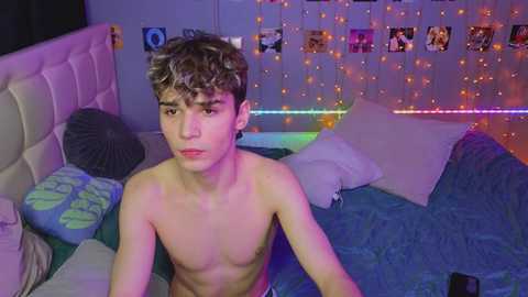 Media: Video of a young, shirtless, slim Caucasian boy with curly brown hair, sitting on a bed with a blue blanket, surrounded by colorful fairy lights and framed photos.