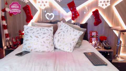 Media: Video of a cozy, festive bedroom with white duvet, pillows, and a red TV remote on the bed. Decorated with red stockings, white snowflakes, and a heart-shaped light.