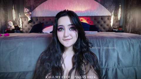 Media: Video of a young woman with long, wavy black hair, wearing dramatic makeup, sitting on a bed with a grey duvet and tufted headboard. Bedroom background features plush pillows and a pink stuffed animal. Text overlays her face.